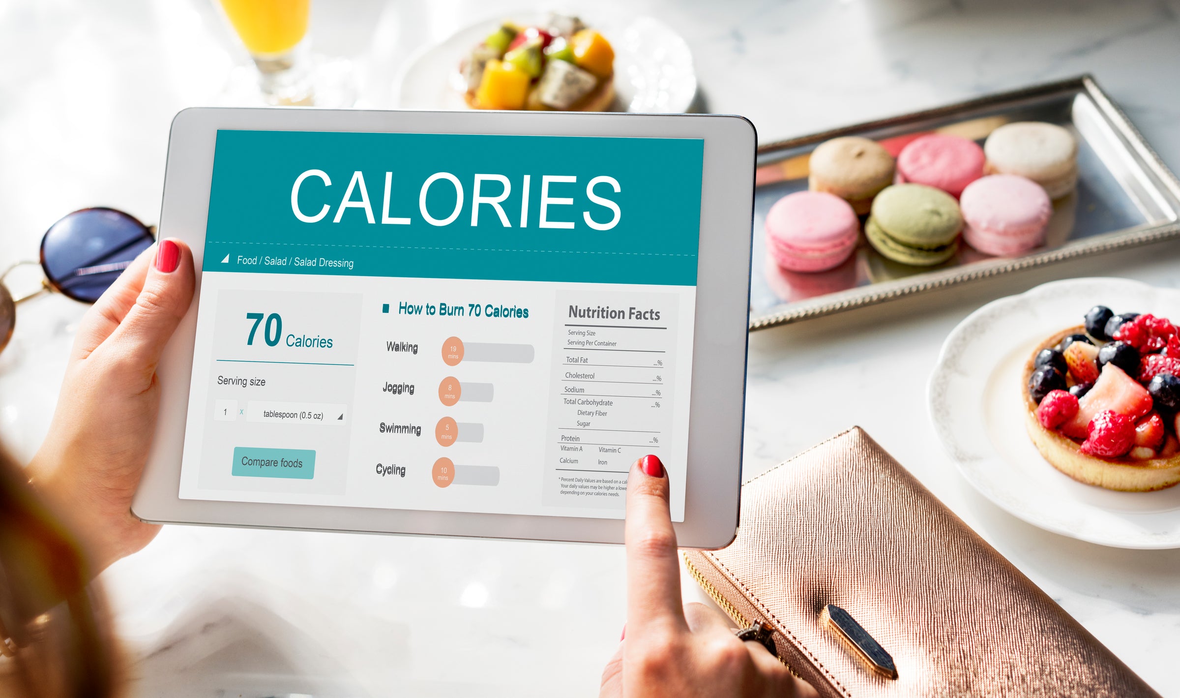 Seorang memegang tablet yang menampilkan informasi kalori dan cara membakar 70 kalori, dengan latar makanan manis seperti macaron dan tart buah di atas meja.