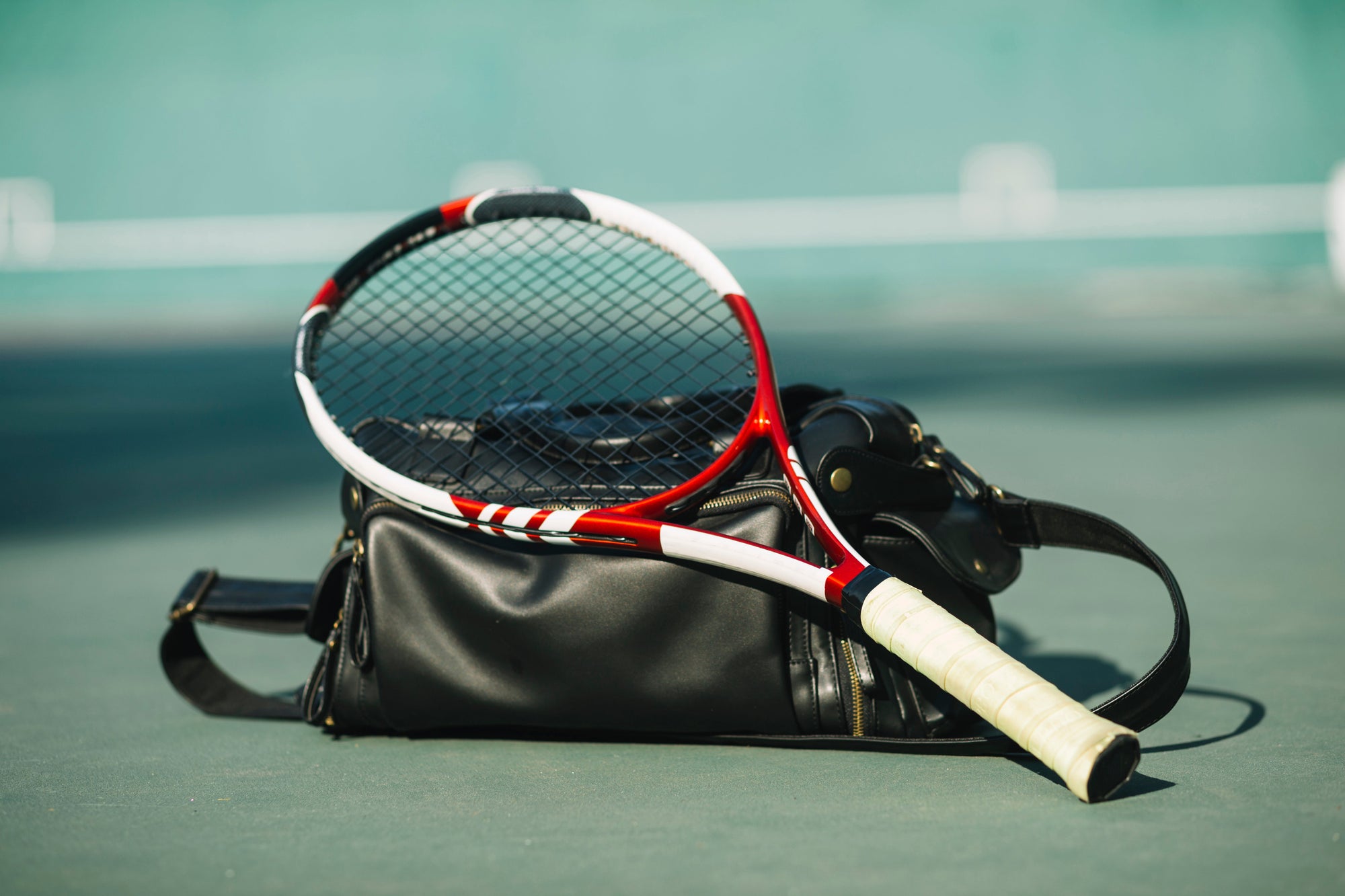 Sebuah raket tenis berwarna merah-putih diletakkan di atas tas olahraga hitam di lapangan tenis luar ruangan.