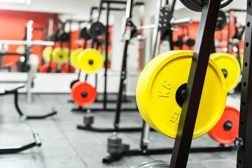 Piringan beban gym berwarna kuning dan merah tersusun di rak, berada di dalam ruang latihan beban modern.