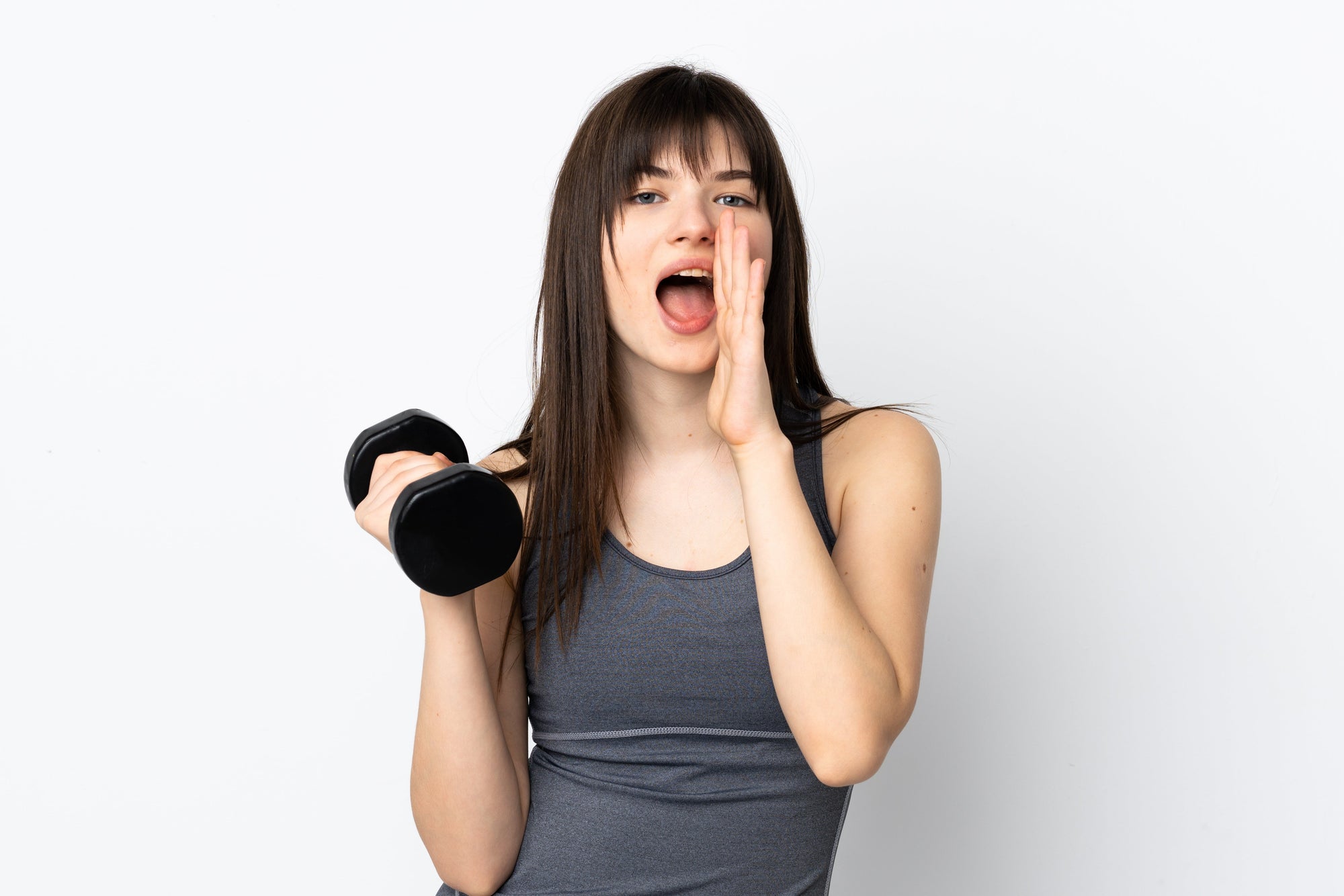 Wanita muda mengenakan tank top olahraga memegang dumbbell hitam sambil berteriak atau memanggil dengan tangan di dekat mulut, berdiri di depan latar belakang putih