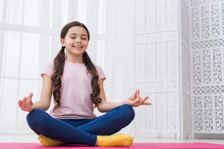 Anak perempuan melakukan yoga dengan pose meditasi duduk di atas matras berwarna pink sambil tersenyum