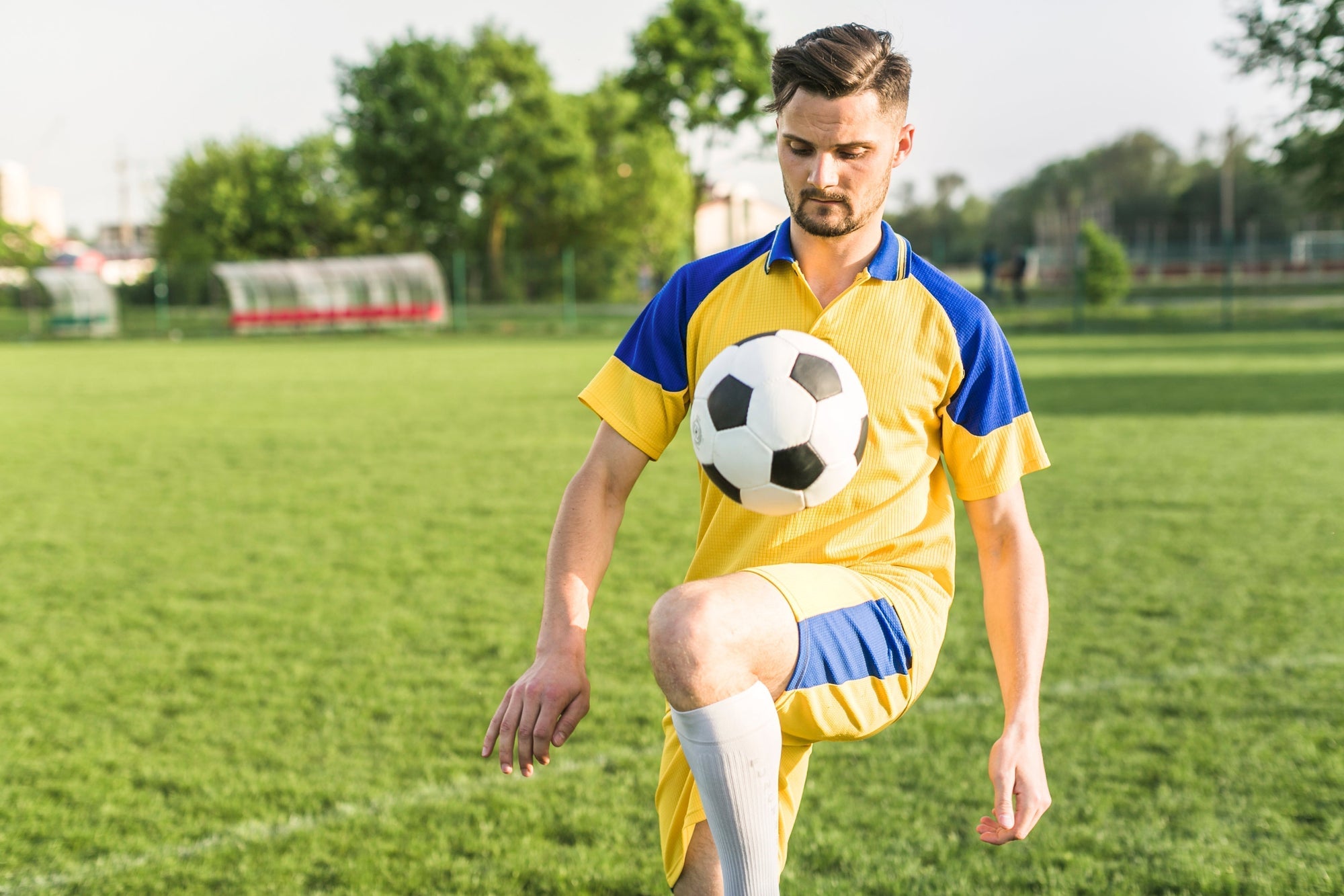 Seorang pemain sepak bola mengenakan jersey kuning dan biru sedang melakukan juggling bola di lapangan rumput terbuka pada siang hari.