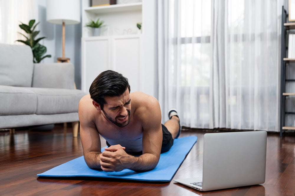 Pria berolahraga di rumah di atas matras biru sambil mengikuti panduan dari laptop, tampak kesakitan saat melakukan plank di ruang tamu yang terang.