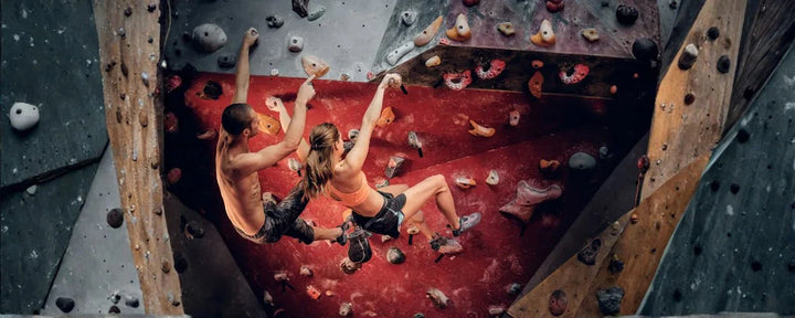 Dua orang sedang memanjat dinding bouldering dengan fokus dan kekuatan, menggambarkan tantangan climbing vs gym yang melatih otot sekaligus ketangkasan