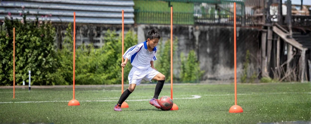 seorang anak yang berlatih menggiring bola menggunakan cone dan tiang koordinasi, dua perlengkapan penting dalam daftar peralatan latihan sepakbola