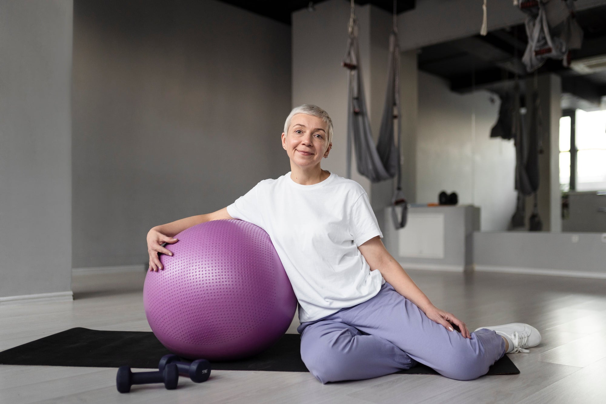 Seorang wanita duduk di gym memeluk bola yoga ungu, menunjukkan manfaat bola yoga untuk core, keseimbangan, dan fleksibilitas.