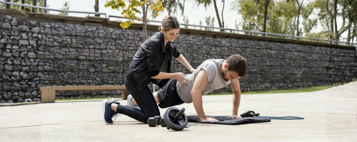 Seorang pria sedang melakukan push-up di luar ruangan dengan bantuan seorang pelatih wanita sebagai salah satu rangkaian gerakan circuit training