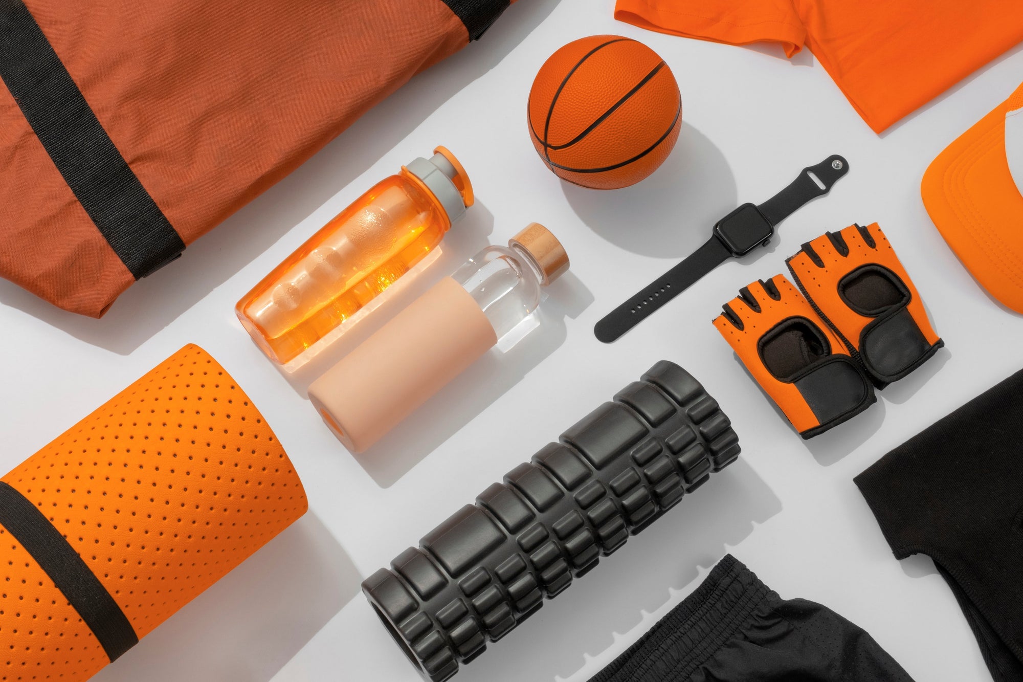 Flat lay perlengkapan olahraga berwarna oranye dan hitam, termasuk matras yoga, foam roller, botol minum, sarung tangan gym, smartwatch, dan bola basket.