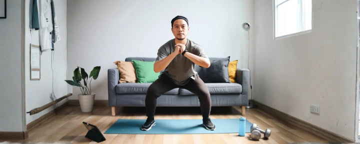 seorang pria muda berbaju abu-abu sedang melakukan latihan squat sebagai upaya untuk mengikuti jadwal workout di rumah agar tubuh bugar