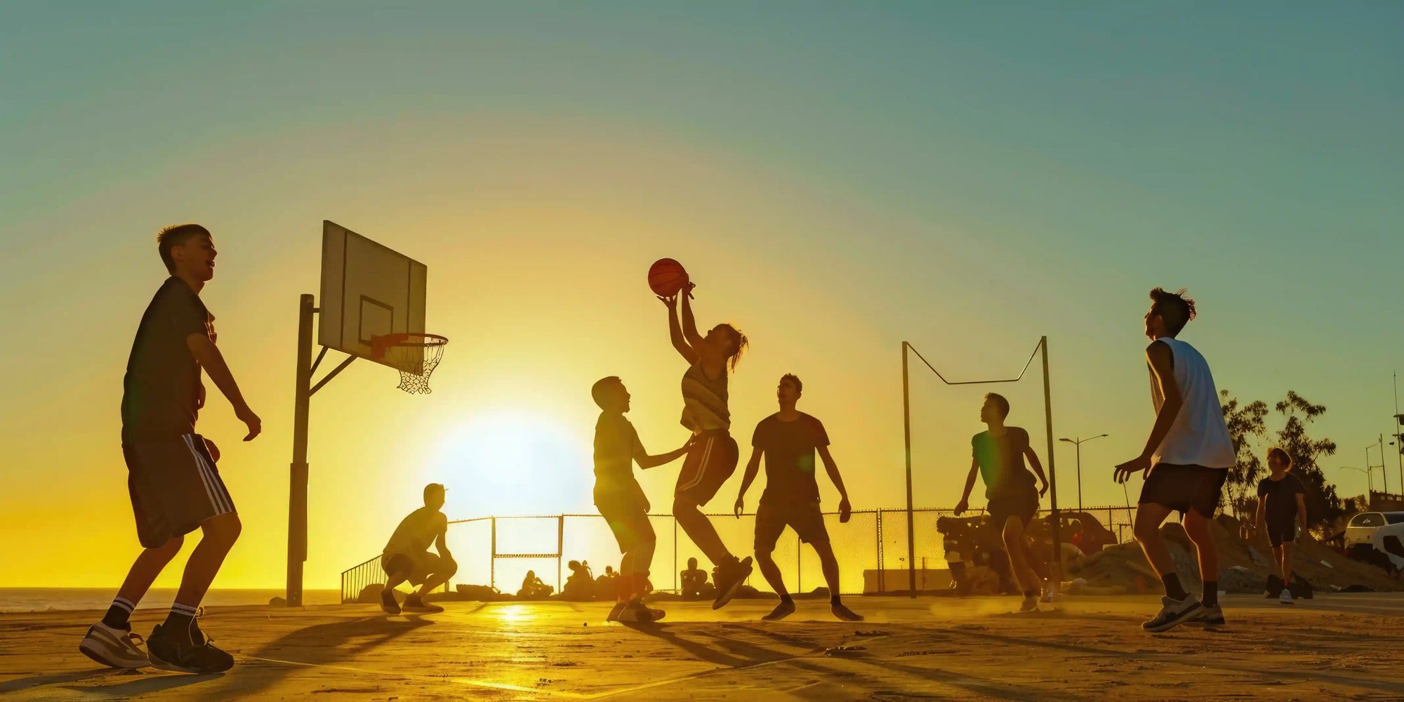 sekelompok orang bermain basket di lapangan outdoor saat matahari terbenam, menggambarkan manfaat bermain bola dalam meningkatkan kebugaran, kerja sama tim, dan kesehatan mental