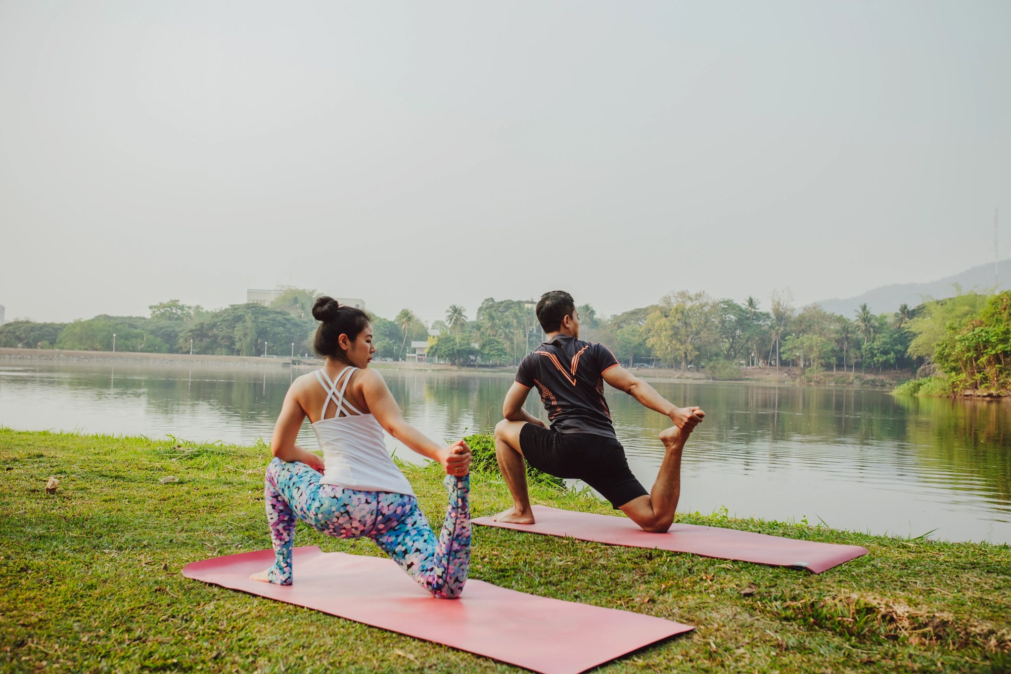 Pasangan sedang berolahraga di tepi danau dengan matras yoga, menggambarkan manfaat senam pagi untuk tubuh lebih bugar dan rileks