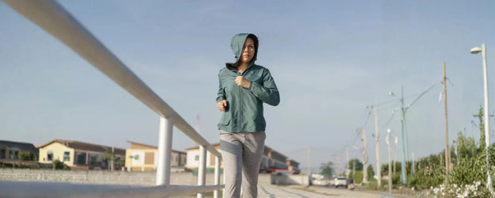 Seorang wanita berlari mengenakan outfit lari pagi berupa jaket hoodie hijau dan celana training abu-abu, tampil sporty dan penuh semangat