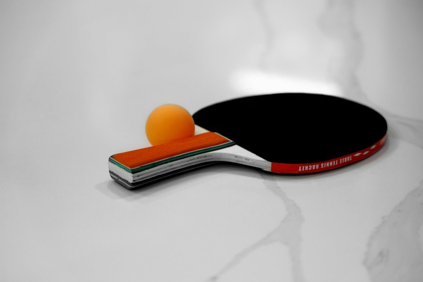 Bet tenis meja berwarna hitam dengan bola pingpong oranye di atas meja putih polos