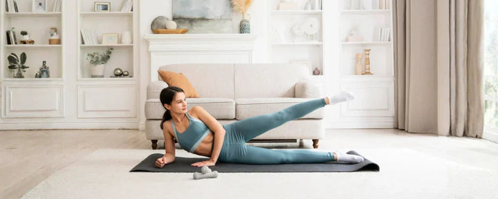 Seorang wanita melakukan gerakan pilates di rumah di atas matras yoga, mengenakan pakaian olahraga berwarna putih dan abu-abu.