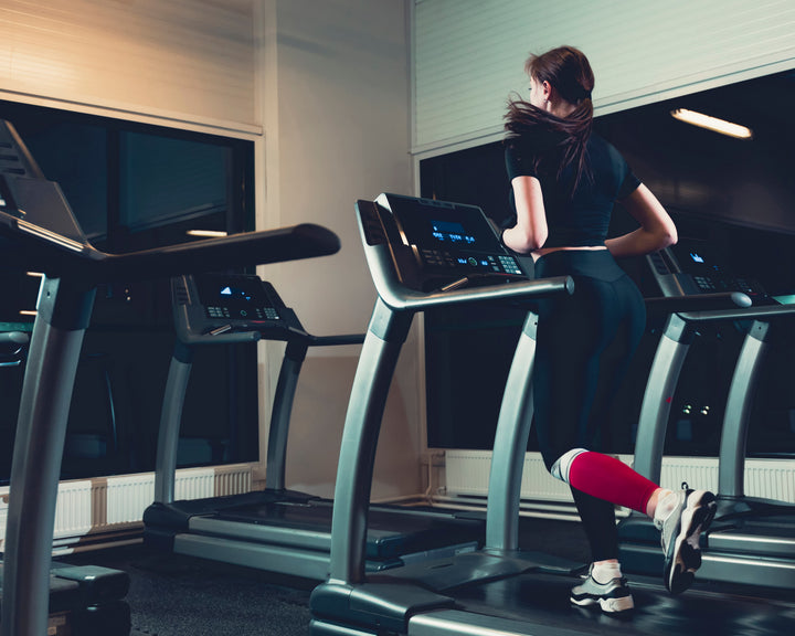 Perempuan berlari di atas treadmill di dalam gym modern saat malam hari, mengenakan pakaian olahraga warna hitam dan legging merah.