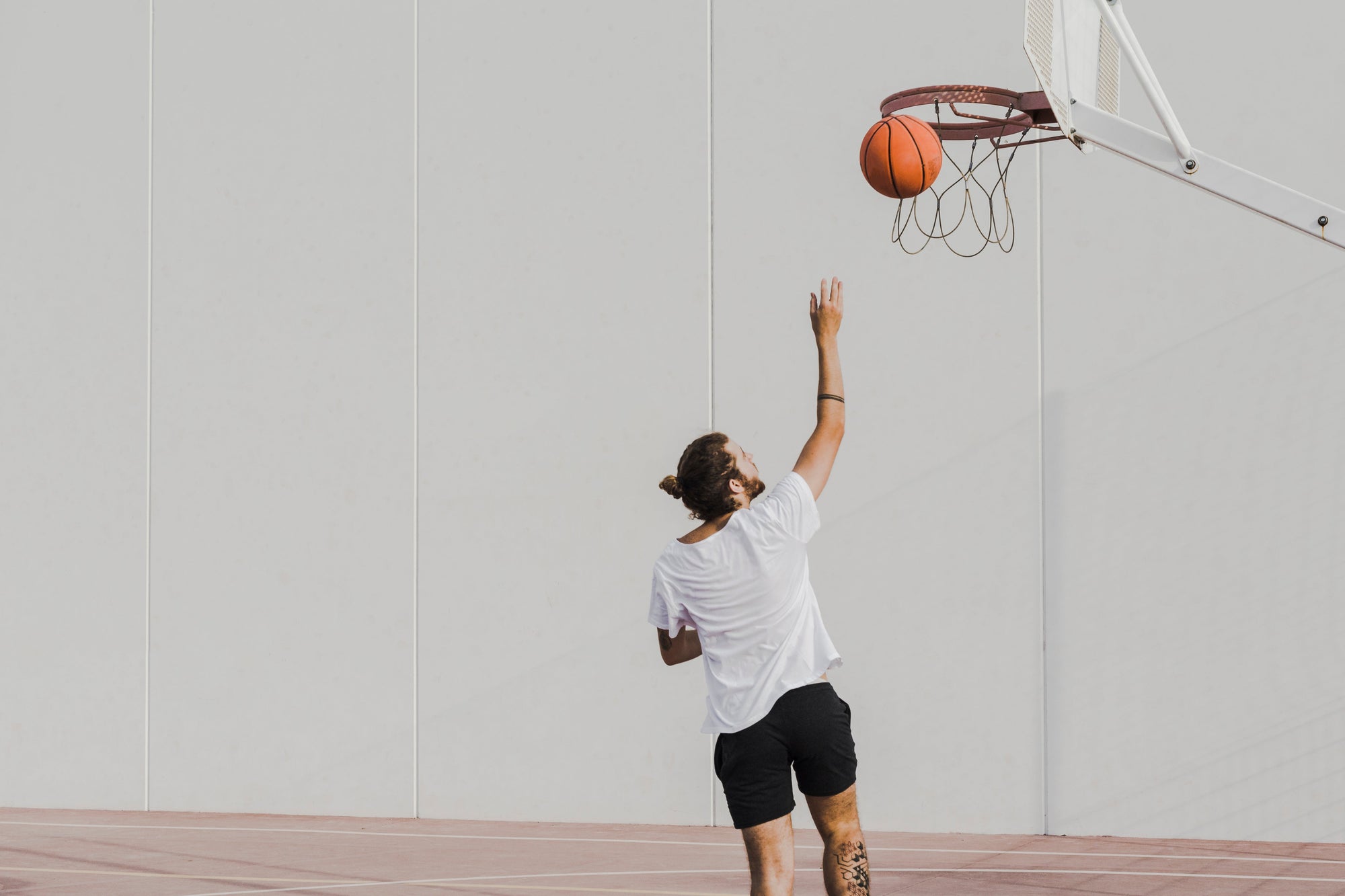 seorang pria muda yang sedang melakukan tembakan lay-up ke arah ring basket di lapangan luar ruangan, mengenakan kaos putih longgar dan celana pendek hitam.
