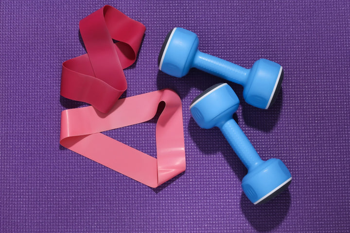 Foto close-up resistance bands vs dumbbell di atas matras ungu, memperlihatkan dua pilihan alat latihan populer