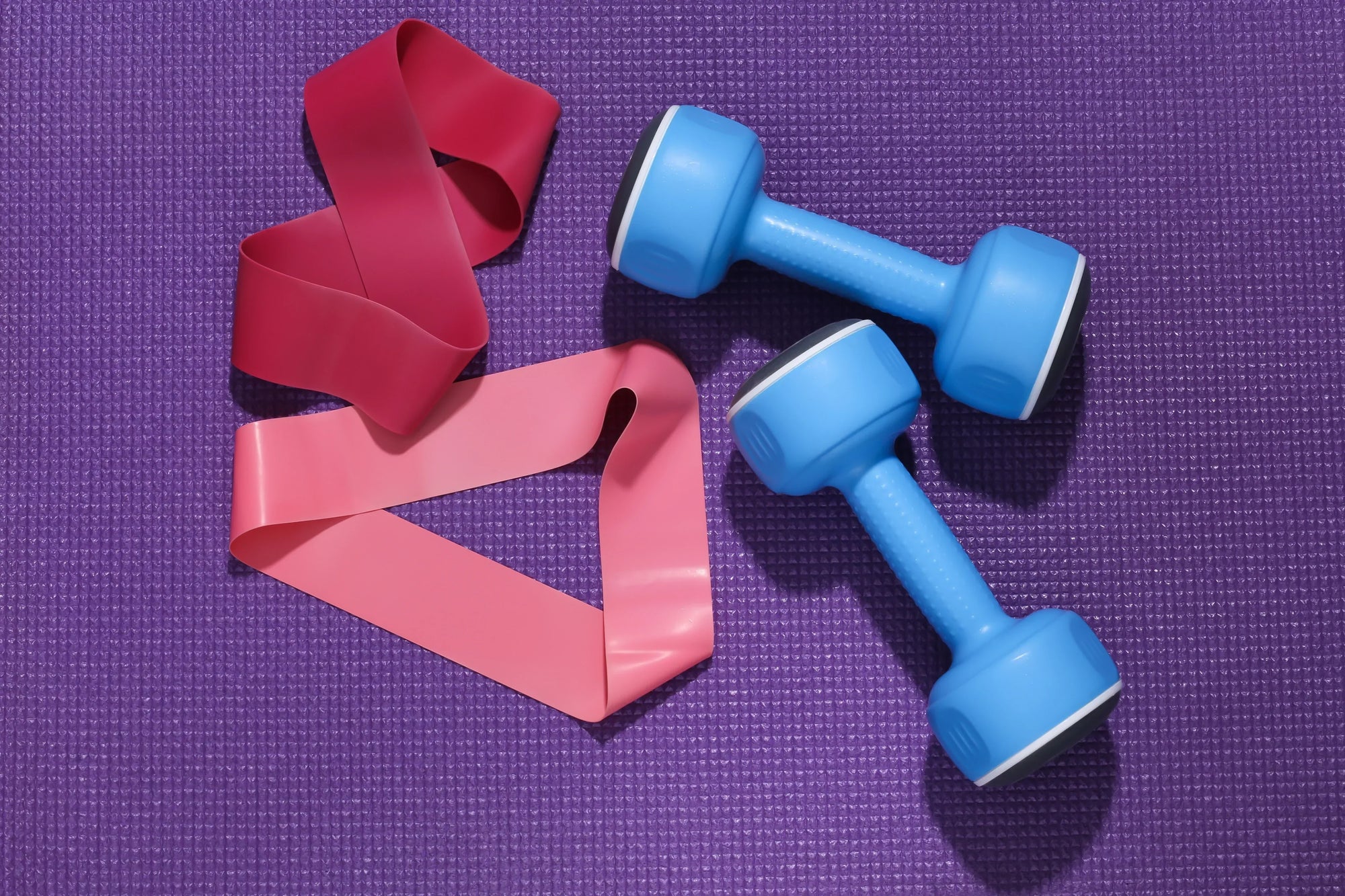 Foto close-up resistance bands vs dumbbell di atas matras ungu, memperlihatkan dua pilihan alat latihan populer