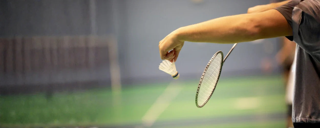 seorang pemain yang sedang melakukan servis bulu tangkis, dengan shuttlecock dipegang rendah dan raket siap mengayun untuk memulai permainan