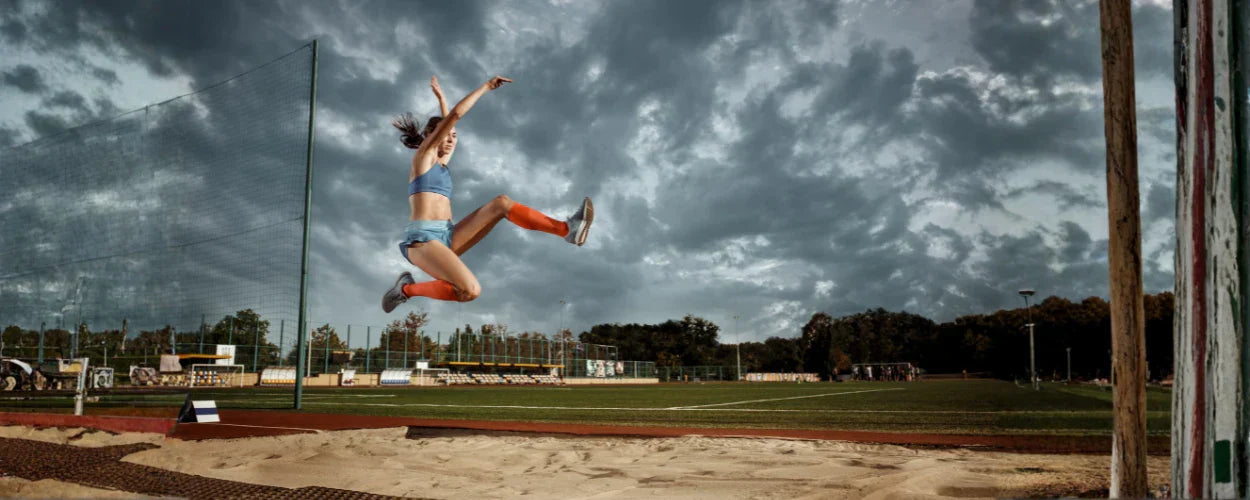 Seorang atlet tampak melayang di udara saat mendarat di bak pasir, menggambarkan teknik dasar lompat jauh dengan gerakan yang kuat dan terukur