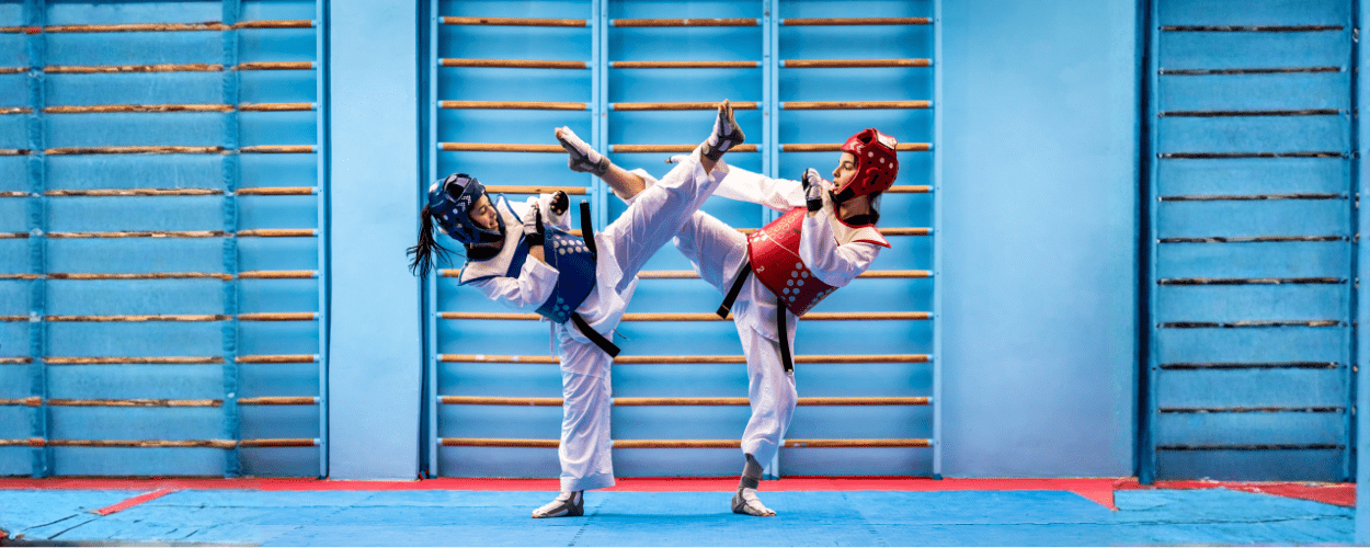 dua atlet sedang mempraktikkan teknik dasar taekwondo berupa tendangan tinggi dengan perlengkapan pelindung lengkap di dalam dojo