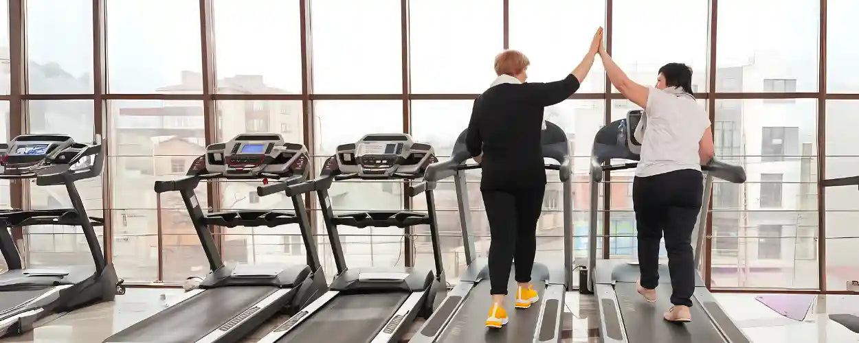 Dua wanita bersemangat berlatih bersama di gym menggunakan treadmill untuk menurunkan berat badan secara sehat dan menyenangkan