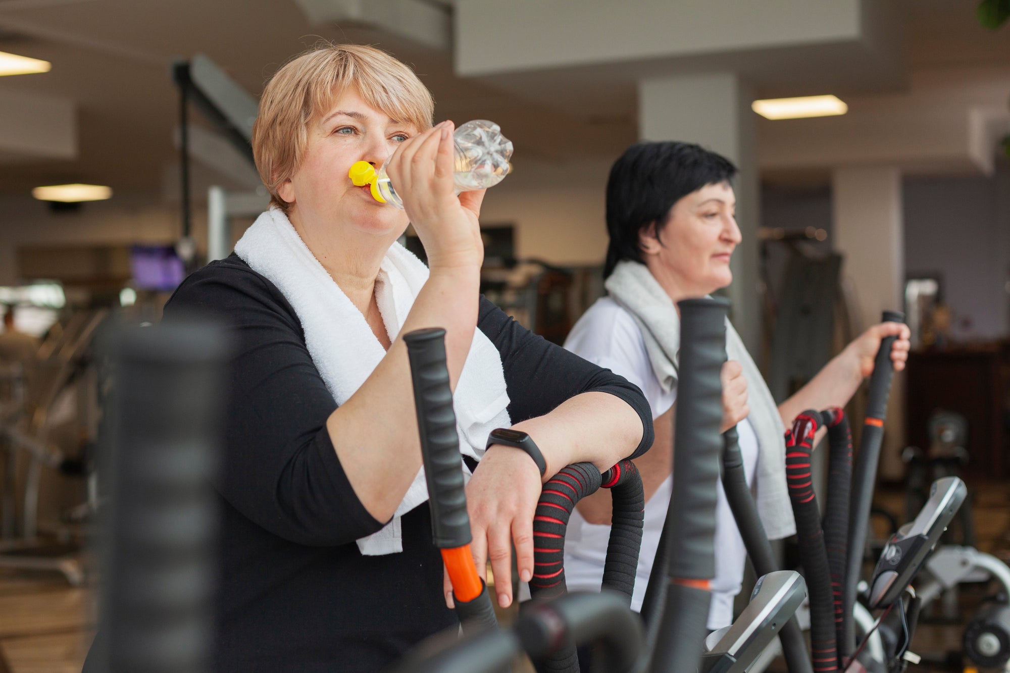 Dua wanita dewasa berolahraga di gym menggunakan mesin elliptical, salah satunya sedang minum air dan mengenakan handuk di leher sebagai bagian dari rutinitas kardio.