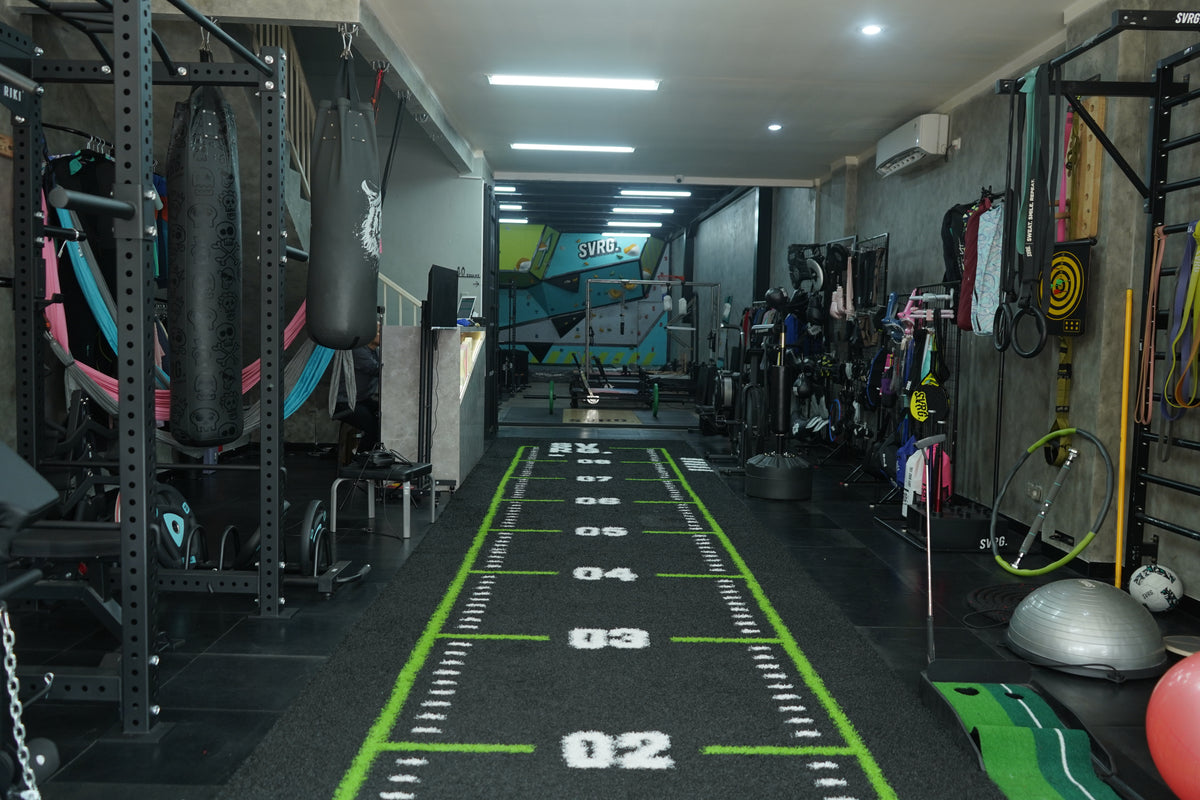 Interior studio gym SVRG dengan nuansa industrial, dilengkapi alat-alat fungsional seperti punching bag, power rack, resistance band, yoga ball, dan turf track dengan angka jarak untuk latihan sled push dan agility.